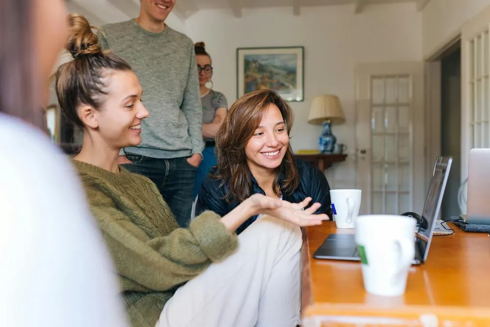 A group of smiling young adults gathered around a laptop at a wooden table in a bright room.