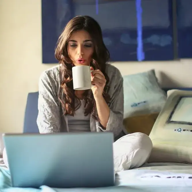 A woman sitting on a bed with a laptop, holding a white mug to her face in a cozy room.